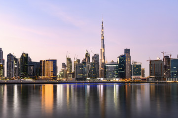 Obraz premium Panoramic view of the illuminated Dubai skyline during sunset with the magnificent Burj Khalifa and many others skyscrapers reflected on a silky smooth water flowing in the foreground. Dubai.
