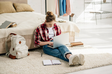 Teenager with laptop and books sitting on carpet and doing homework