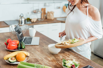Cropped view of cheerful girl holding chopping board with cut bananas and avocados