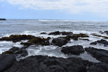 Fototapeta na wymiar Waves Crashing on Rocky Shore in Hawaii white foam as the waves wash over the rocks under a blue sky