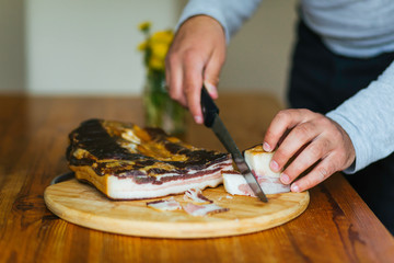 Young man cutting bacon on a wooden cutting board with a knife on a wooden table in the kitchen. 