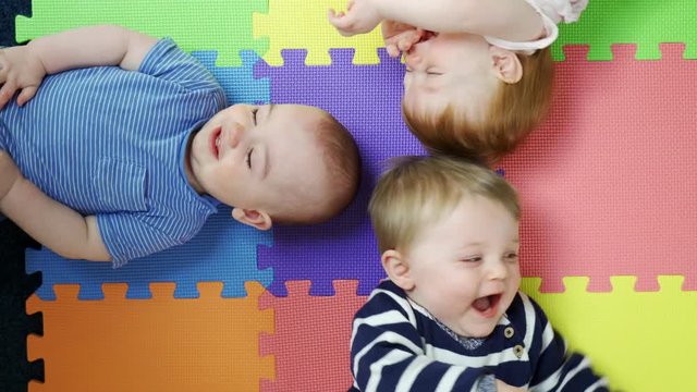 Overhead View Of Babies Lying On Mat At Nursery PlaygroupBeing Tickled By Mothers And Laughing