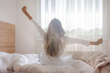 Young woman waking up in her bedroom, sitting on the bed stretching arms by the window - obrazy, fototapety, plakaty