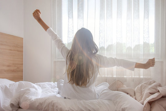 Young woman waking up in her bedroom, sitting on the bed stretching arms by the window