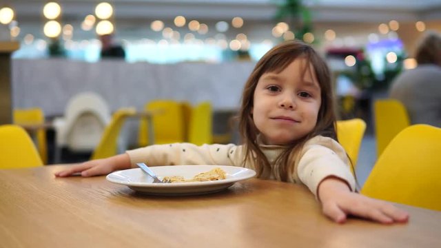 Blonde girl 4-5 years old eats pancake. Child on the food court. Eating concept. In the background a blurred defocused image of people bokeh light bulbs.