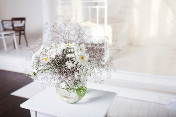 Glass vase  white camomile floweers  in light cozy bedroom interior. White wall, retro bed, sunlight from window, copy space