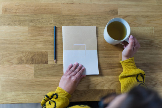 L&aacute;pis, m&atilde;o de mulher em posi&ccedil;&atilde;o de abrir um caderno fechado e a outra m&atilde;o segurando uma caneca branca com ch&aacute;. Numa bancada de madeira.