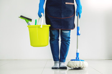 Young housekeeper cleaning floor mobbing holding mop and plastic bucket with brushes, gloves and...