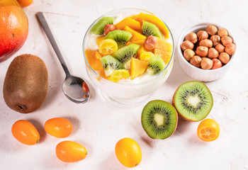 Glass bowl with yogurt, and fruits, metallic spoon and bowl of nuts on whitetable. 