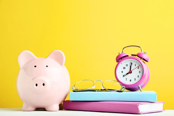 Pink piggy bank with alarm clock, glasses and books on yellow background