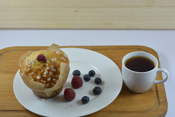 a morning jam muffin on a white plate next to raspberries and blueberries on a wooden chopping board next to a white cup of coffee