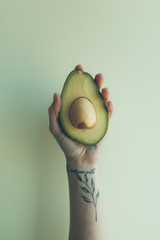 a female hand with a branch-shaped tattoo holds a half of a raw avocado on a plain background....