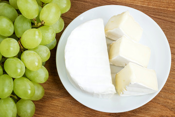 White plate with pieces of tasty cheese camembert and sweet green grapes on a brown wooden cutting board. Soft cheese covered with edible white mold.