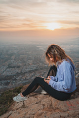 Young woman sitting and using the mobile on the top of the mountain looking at the city from above, during nice sunset