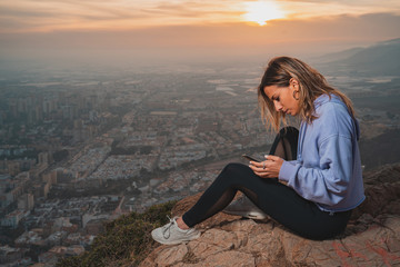 Naklejka na ściany i meble Young woman sitting and using the mobile on the top of the mountain looking at the city from above, during nice sunset