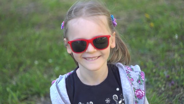 Blonde European happy Child (6 years old girl) with two tails on a Sunny green meadow laughs, talks, takes off the red sunglasses, close-up