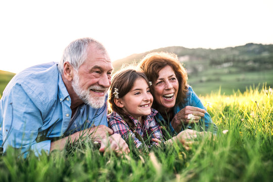 Senior Couple With Granddaughter Outside In Spring Nature, Relaxing On The Grass.