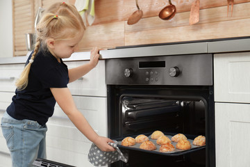 Little girl taking out baking sheet with cookies from oven in kitchen