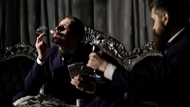 Luxury men sitting on sofa and smoking cigars in men's club and solving global problems. Collusion in business and global conspiracy between two successful businessmen. Business, money concept.