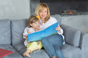 Mother and little daughter are playing, reading a book, sitting and enjoying in a sofa.