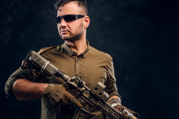 Fototapeta na wymiar Portrait of a stylish man wearing shirt sunglasses holding assault rifle and looking sideways.