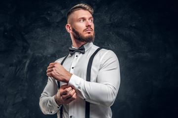 Stylishly dressed young man in shirt with bow tie and suspenders. Studio photo against dark wall background