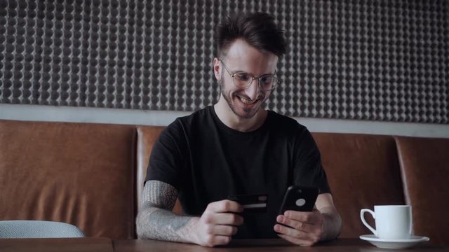 Young smily guy in a glasses, shops online through his smart phone. Holding creadit cart and the smart phone