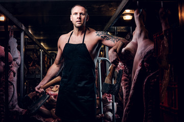 Fototapeta na wymiar Shirtless tattooed butcher in apron posing with ax in a refrigerated warehouse in the midst of meat carcasses