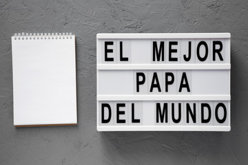 'El Mejor Papa Del Mundo' words on lightbox, blank notepad over gray background. Overhead, from above, flat lay. Father's day. Copy space.
