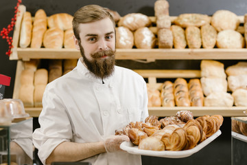 Fototapeta na wymiar Charismatic baker with a beard and mustache stands with a tray with fresh pastries on the background of shelves with fresh bread in the bakery