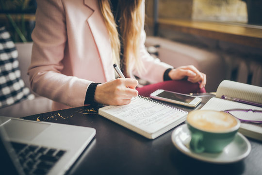 Close Up Caucasian Woman Hand On Wooden Table Inside Cafe Makes Notes In Notebook. Subject Freelancer Blogger Journalist At Work. Unrecognizable Person. Technology Phone And Laptop With Cup Coffee