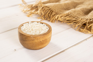 Fototapeta na wymiar Lot of whole white jasmine rice grains in a wooden bowl on jute cloth on white wood