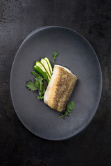 Fried Danish skrei cod fish filet with baby zucchini and lettuce as top view on a modern design plate
