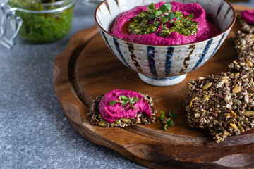 Fototapeta na wymiar Roasted beet hummus in ceramic bowl, flax seed crackers on wooden cutting board, pesto sauce jar - vegetarian, vegan food concept