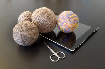 Hobby on the Internet. Gray and colored wool yarns for home knitting, a tablet computer and small scissors lie on a dark wooden table.
