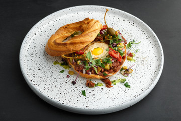 bagel filled with beef tartare, hummus, quail egg yolk, pickled capers, microgreens, dried tomatoes, mustard and cutted onions. Served on white textured plate.