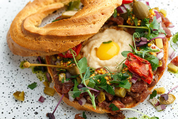 bagel filled with beef tartare, hummus, quail egg yolk, pickled capers, microgreens, dried tomatoes, mustard and cutted onions. Served on white textured plate. Close up view