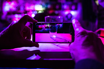 A person is taking a photograph with his phone to a glass in a pub. The glass is Illuminated by colored lights and the background is out of focus. Horizontal photo