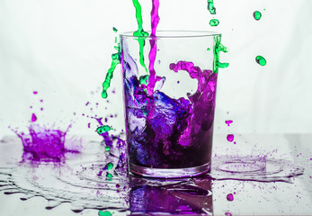 Green and purple liquid spilling into and outside a glass standing on a glass floor.