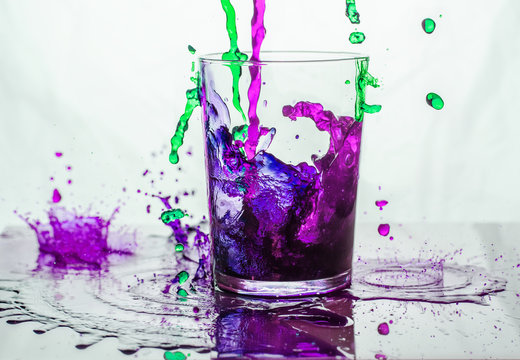 Green And Purple Liquid Spilling Into And Outside A Glass Standing On A Glass Floor.