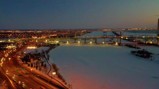 A slow moving aerial shot at twilight that flies over a half-frozen river, showing a long bridge and a brightly city in the background.