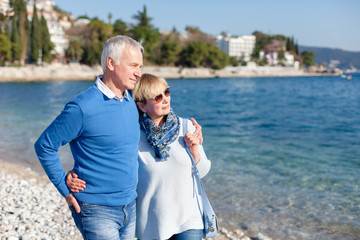 Senior couple is hugging and walking at sea beach outdoor. Happy man and woman are relaxing, traveling, enjoying life and retirement. Concept of wellbeing, family love, care.