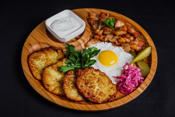 Potato pancakes with a fried egg, bacon, sourcream, horse radish, pickles and parsley on a wooden plate on black background