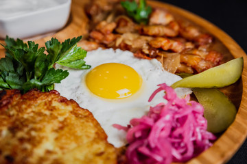 Potato pancakes with a fried egg, bacon, sourcream, horse radish, pickles and parsley on a wooden plate on black background