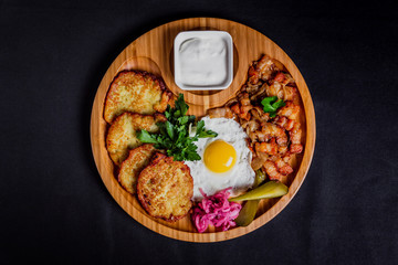 Potato pancakes with a fried egg, bacon, sourcream, horse radish, pickles and parsley on a wooden plate on black background