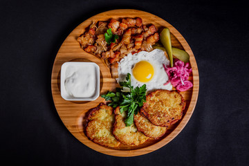 Potato pancakes with a fried egg, bacon, sourcream, horse radish, pickles and parsley on a wooden plate on black background