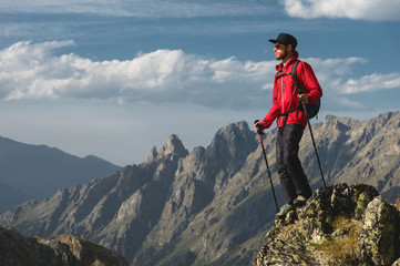 Porter of a bearded traveler hipster in sunglasses and a cap with a backpack and trekking poles stands on a high cliff near the cliff on the background of epic mountains of rocks and clouds