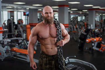 Fototapeta na wymiar A bald, bearded bodybuilder with large relief muscles holds a heavy thick chain in his hands. Bright gym.