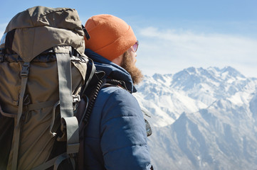 Portrait of a bearded man in sunglasses and a warm jacket with a backpack on his back against the background of snowy mountains on a sunny day. The concept of tourism and travel