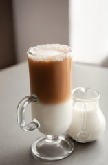 Glass cup of tasty frappe coffee and jug of milk on grey table